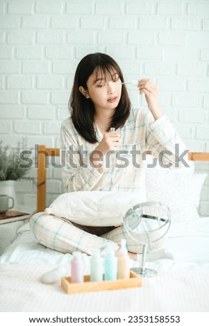 Skincare Concept. Beautiful Asian Woman Applying Face Serum With Dropper While Sitting In Front Of Mirror At Home, Smiling Asian Woman Moisturizing Skin, Enjoying Self-Care Routine