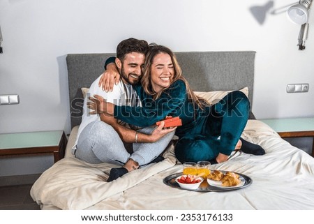 Boy in pajamas gives a gift to his wife as they have breakfast in bed in the bedroom of their modern home. Very happy wife hugging her husband. Anniversary celebration. Romantic breakfast
