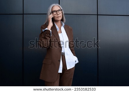 60 year old successful slender gray-haired business woman dressed stylishly speaks on the phone while walking along the wall of the business center Royalty-Free Stock Photo #2353182533