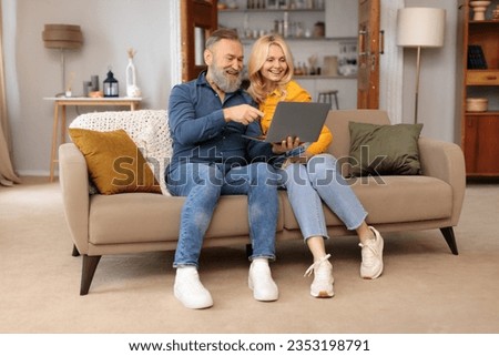 Full Length Of Romantic Mature Spouses Relaxing With Laptop At Home, Man Pointing At Computer, Websurfing And Having Fun Online. Older Spouses Enjoying Gadget Use And Internet Technology
