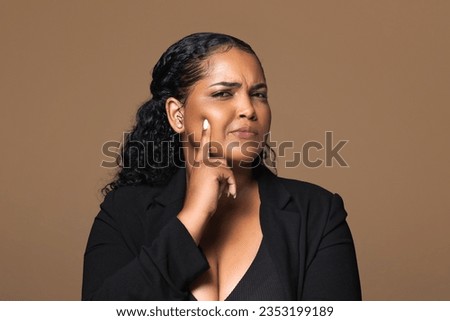 Pensive brazilian curly woman thinking, touching cheek and looking at camera isolated on brown background, studio shot. Idea creating, brainstorm, ad and offer, fashion blog and lifestyle