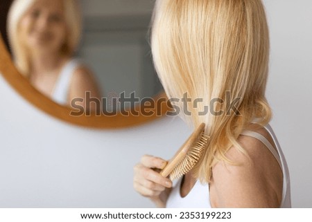 Rear view of middle aged woman brushing hair with wooden hairbrush looking at mirror in bathroom at home. Unrecognizable mature female combing blonde hair Indoor