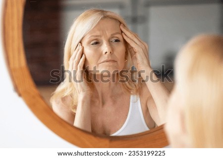 Upset middle aged woman looking at mirror and touching wrinkles on her face, lady examining fine lines on forehead, suffering skin aging, selective focus on reflection Royalty-Free Stock Photo #2353199225