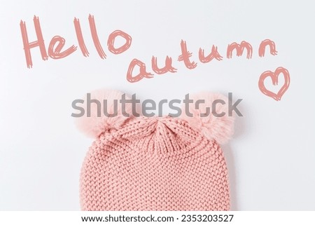 Pink knitted hat with pumpons and the inscription hello autumn with a heart. Warm children's hat on a white background, top view