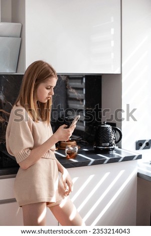 Teen girl using smartphone in kitchen. Remote education, online courses for teens, online shopping, good morning concept. Neutral colors. Vertical image.