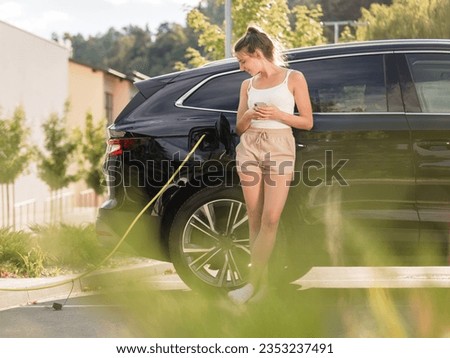 Young woman charging her elegant black EV car at a public electric station in the neighborhood. Clean and alternative energy concept.