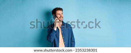 Handsome caucasian man talking on mobile phone, looking aside at empty space with carefree smile, standing on blue background.