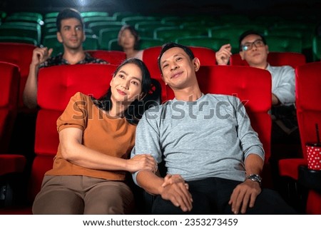 Happy trip of asian couple sweet sit togather on red seat and looking  movie in cinema with fun a popcorn on hand, people relax and holiday activity on a weekend Royalty-Free Stock Photo #2353273459