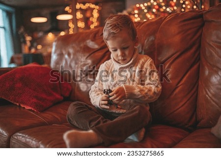 Smile small child being active having fun in motion and dreamer waiting for miracle Santa. Cheerful kid in knitwear outfit playing with toy indulge indoor on sofa eve 25 December garlands lights Noel