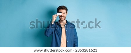 Happy male client holding plastic credit card on eye and smiling, standing in casual outfit on blue background.