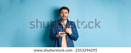 Celebration and holiday concept. Surprised guy holding birthday cake with candle and pointing at himself with disbelief, being congratulated with b-day, blue background.