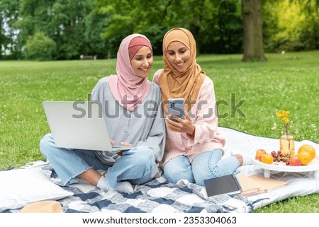 Two beautiful Arab women in a hijab sit in a city park on a picnic, study and work remotely, look at the phone screen, use and type on a laptop. Concept of learning and working outdoors. Recreation.