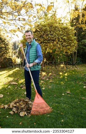 Man raking leaves in garden smiling at camera during fall autumn backyard cleanup Royalty-Free Stock Photo #2353304127