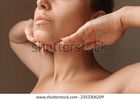 Cropped shot of young caucasian woman touching under the chin with hands massaging her face on dark brown background. Rejuvenation, facelift, facefitness. Exercises from the second chin, pelican neck Royalty-Free Stock Photo #2353306209