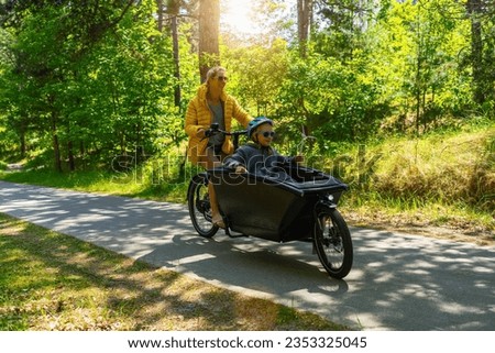Mother and daughter having a ride on road with electric cargo bike holding wind turbine model at summer. Renewable energy concept image.