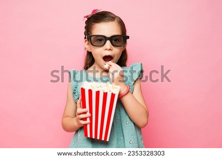 Adorable elementary age kid enjoying eating popcorn while watching a 3d movie with glasses in a pink background