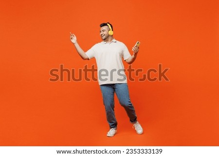 Full body happy young Indian man he wearing white t-shirt casual clothes listen in headphones using mobile cell phone dance isolated on plain orange red background studio portrait. Lifestyle concept