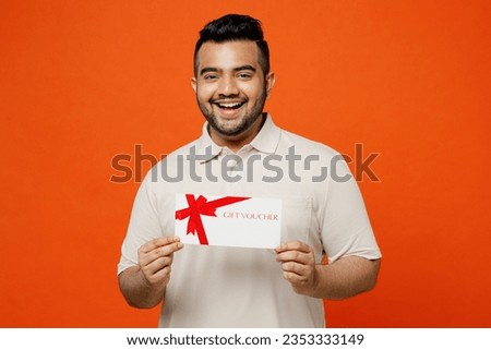 Young smiling happy fun Indian man he wears white t-shirt casual clothes hold gift certificate coupon voucher card for store isolated on plain orange red background studio portrait. Lifestyle concept