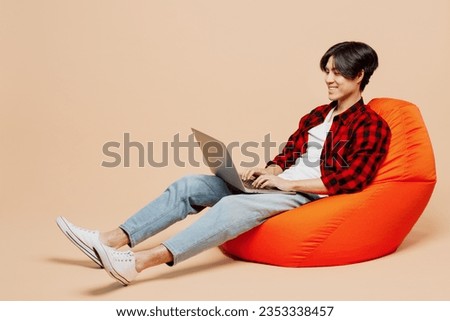 Full body young IT man of Asian ethnicity wear red shirt casual clothes sit in bag chair hold use work on laptop pc computer isolated on plain pastel light beige background studio. Lifestyle concept