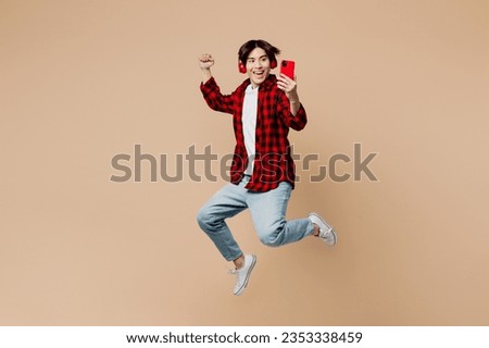 Full body side view young man of Asian ethnicity wear red shirt casual clothes jump high listen to music in headphones use mobile cell phone do winner gesture isolated on plain light beige background