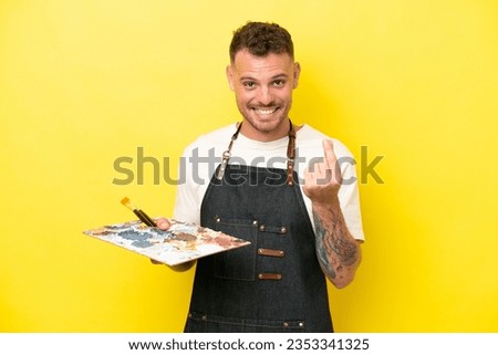 Young artist caucasian man holding a palette isolated on yellow background making money gesture