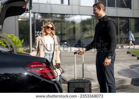 Chauffeur packs a suitcase in a car trunk, businesswoman waiting nearby using luxury taxi service during a business trip. Concept of business transfer services, idea of personal driver. Royalty-Free Stock Photo #2353368073