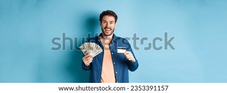 Smiling caucasian guy showing plastic credit card and money, looking happy, standing on blue background.