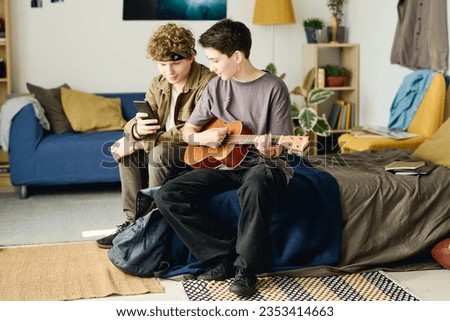 Long shot of two teenagers in casualwear looking at smartphone screen while sitting on bed at home and shooting video for online audience