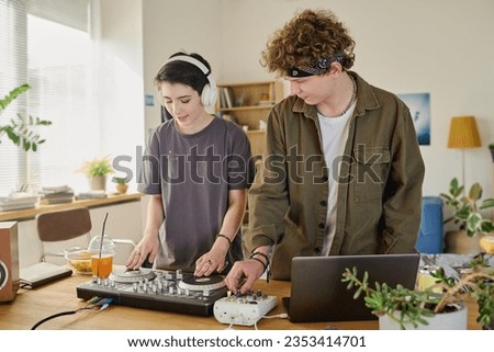 Modern teenage couple creating new computer music and recording it while standing by desk with turntables and laptop in home environment