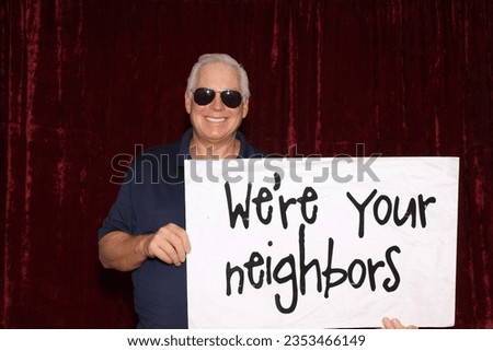 Photo Booth. Large Sign. A man smiles and laughs as he holds a large sign that reads We're your neighbors while in a Photo Booth. Neighbors love a Photo Booth for all parties and events. Photo Booth. 