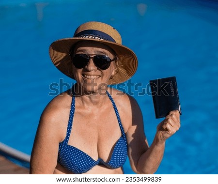 An elderly woman in a hat and sunglasses holds a passport while sitting by the pool. Vacation in retirement. 