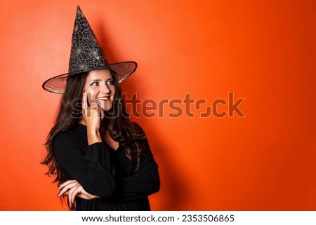 Beautiful Caucasian woman wearing black dress and witch hat portrait on orange background. Copy space. 