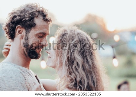 Lifestyle concept of a beautiful young couple in love seperating from their group of friends in the background having an outdoor garden barbecue laughing with wine drinks, outdoors on a sunny day
