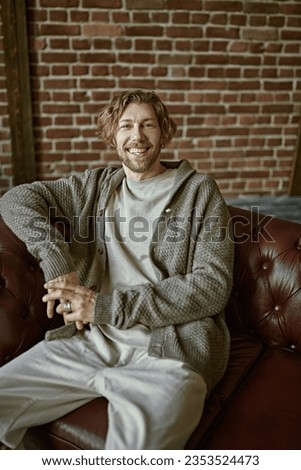 Portrait of a handsome forty-year-old man with curly hair, dressed in a casual knitted cardigan, who looks happily to the camera. Loft interior. Life style. People, emotions. Royalty-Free Stock Photo #2353524473