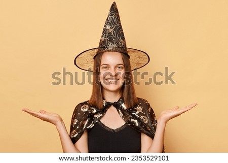 Smiling young woman wearing witch costume and carnival cone hat celebrating halloween isolated over beige background spreading palms presenting copy space for promotion.
