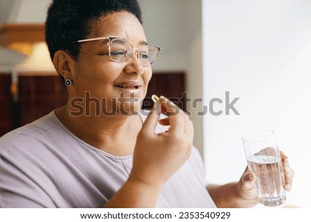 Indoor image of overweight elderly female in glasses taking transparent capsule of fish oil to stay healthy, holding glass of pure water in hand. Food supplements for brain, nerve system support Royalty-Free Stock Photo #2353540929