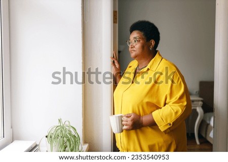 Side view of sad african american overweight female in yellow clothes and glasses standing next to window with cup of hot drink, grandma missing her grandchildren, waiting them to visit her Royalty-Free Stock Photo #2353540953