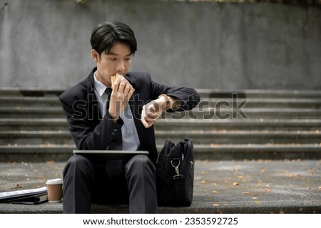 A busy and hungry young Asian businessman or male office worker is checking time on his watch and eating a sandwich for his quick lunch on a staircase in front of the building. Busy urban lifestyle  Royalty-Free Stock Photo #2353592725