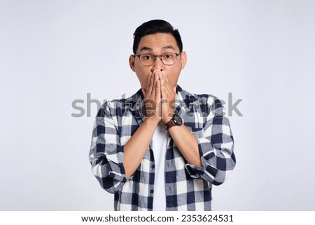Surprised young Asian man in casual shirt covering mouth with hand, looking at camera with amazed facial expression isolated on white background