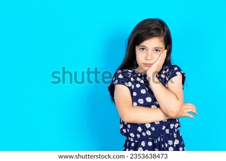 Beautiful kid girl wearing dress over blue background covers eyes with palm and doing stop gesture, tries to hide from everybody.