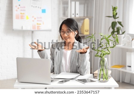Young Asian businesswoman with laptop sitting at table and video chatting in office