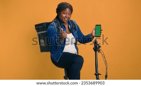 African american female with backpack carries smartphone with isolated green screen display. Friendly courier shows online ordering logistics on cell phone with chromakey mockup template.
