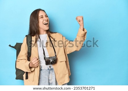Young Caucasian woman traveler with camera and backpack raising fist after a victory, winner concept.