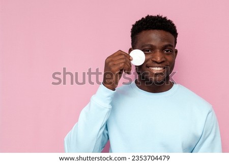 african american man in blue sweater uses cotton pads and smiles on pink isolated background, man takes care of his face skin and wipes it