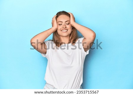 Blonde young woman in white t-shirt on blue background laughs joyfully keeping hands on head. Happiness concept.