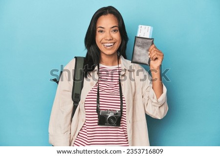 Filipina traveler displaying flight ticket with camera and backpack on blue Royalty-Free Stock Photo #2353716809