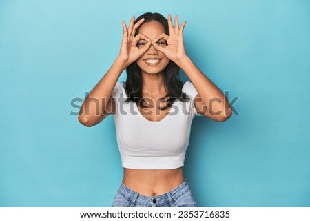 Filipina young woman on blue studio showing okay sign over eyes