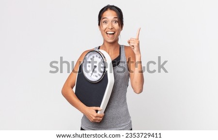 young hispanic woman feeling like a happy and excited genius after realizing an idea and holding a weight scale. diet concept Royalty-Free Stock Photo #2353729411
