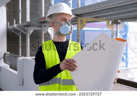 young male builder engineer, architect in hardhat, protective respirator during inspection in construction site, looks at project plan on paper, evaluates, concept of monitoring of structures Royalty-Free Stock Photo #2353729727