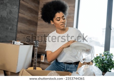 african american woman unpacking tableware near carton packages in new house Royalty-Free Stock Photo #2353744361